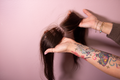 Person with tattooed arm holding a person's hair against a pink background