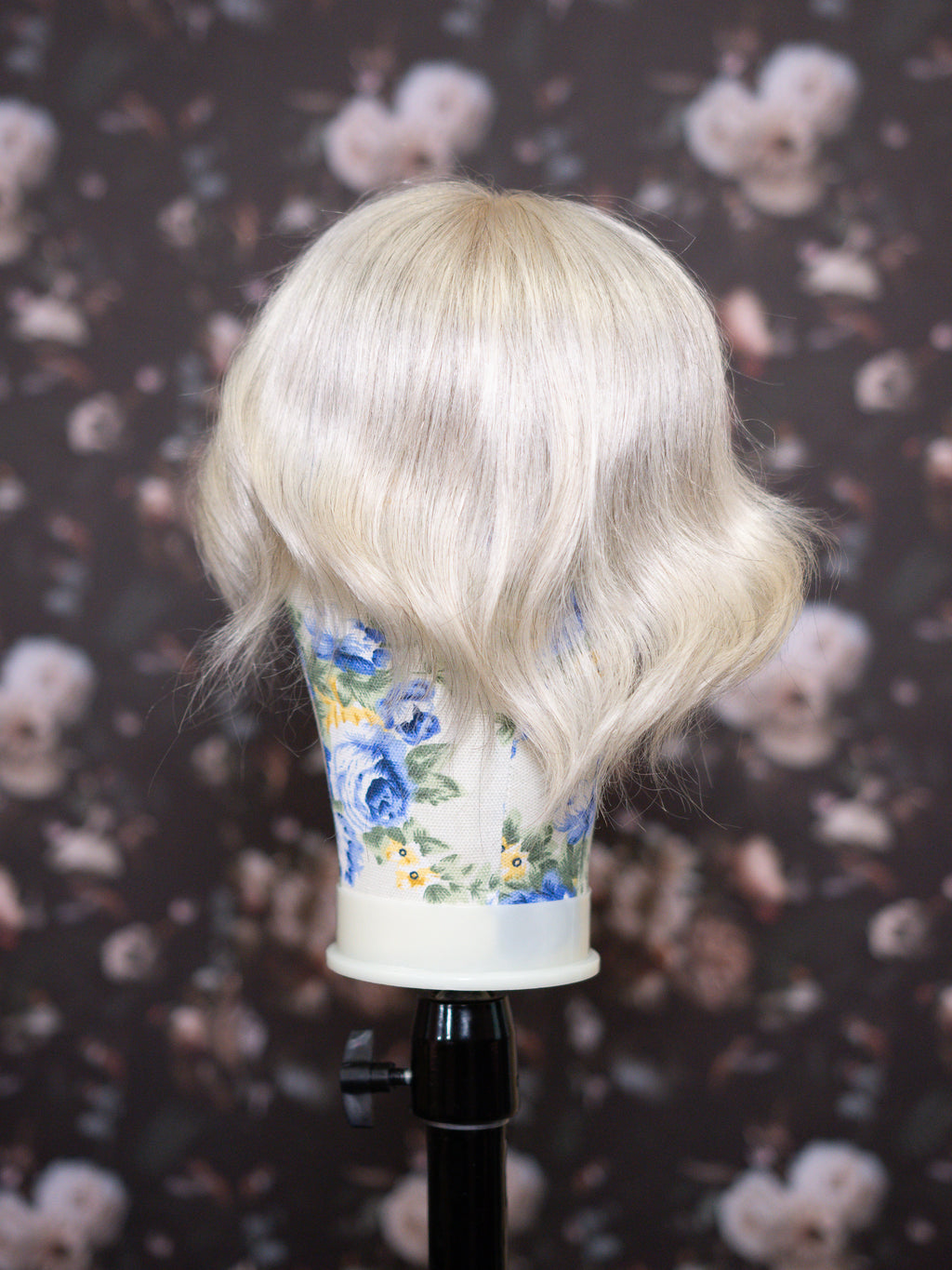 Blonde wig on a mannequin head with a floral-patterned cup against a dark floral background