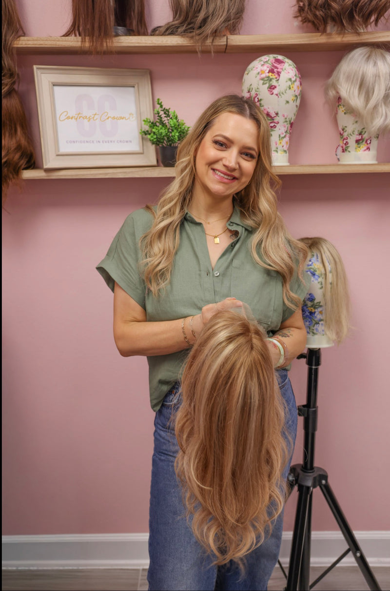 Woman holding a blonde wig in a hair salon setting with shelves of wigs and decor.