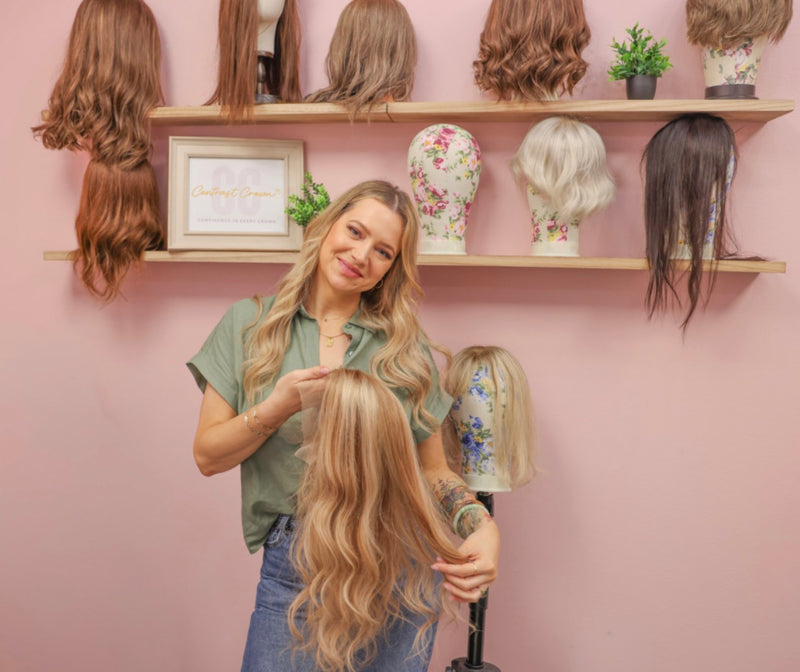 Woman styling a wig with a shelf of wigs and decorative items in the background.