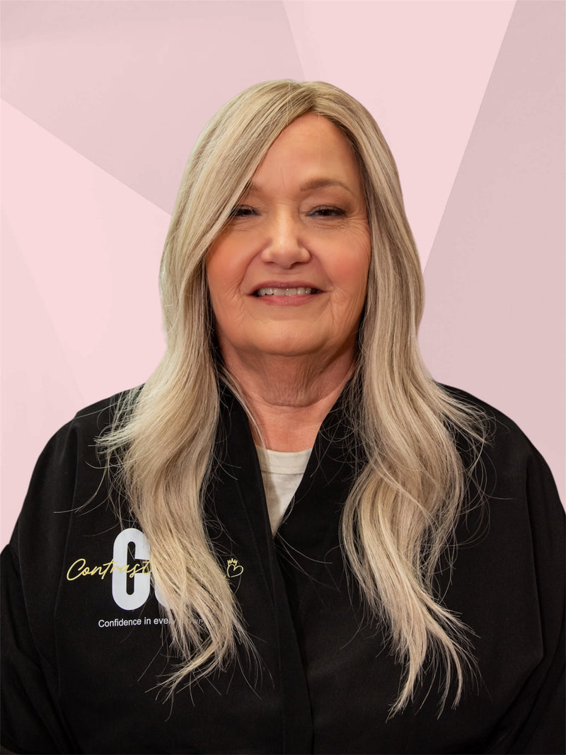 Person with long blonde hair wearing a black jacket with visible branding on a pink background